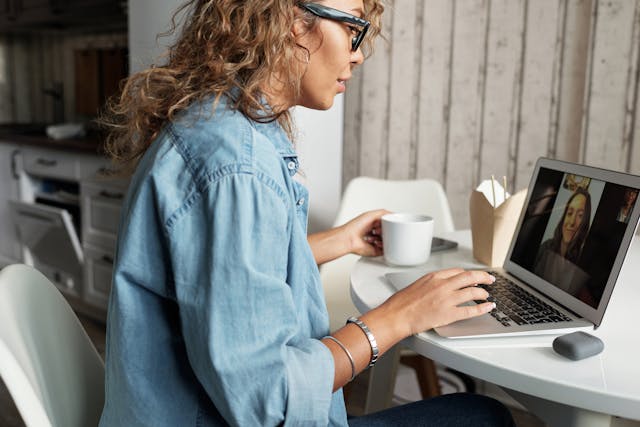 A person sitting at a table with a mug in one hand and using a laptop for a video call with another person